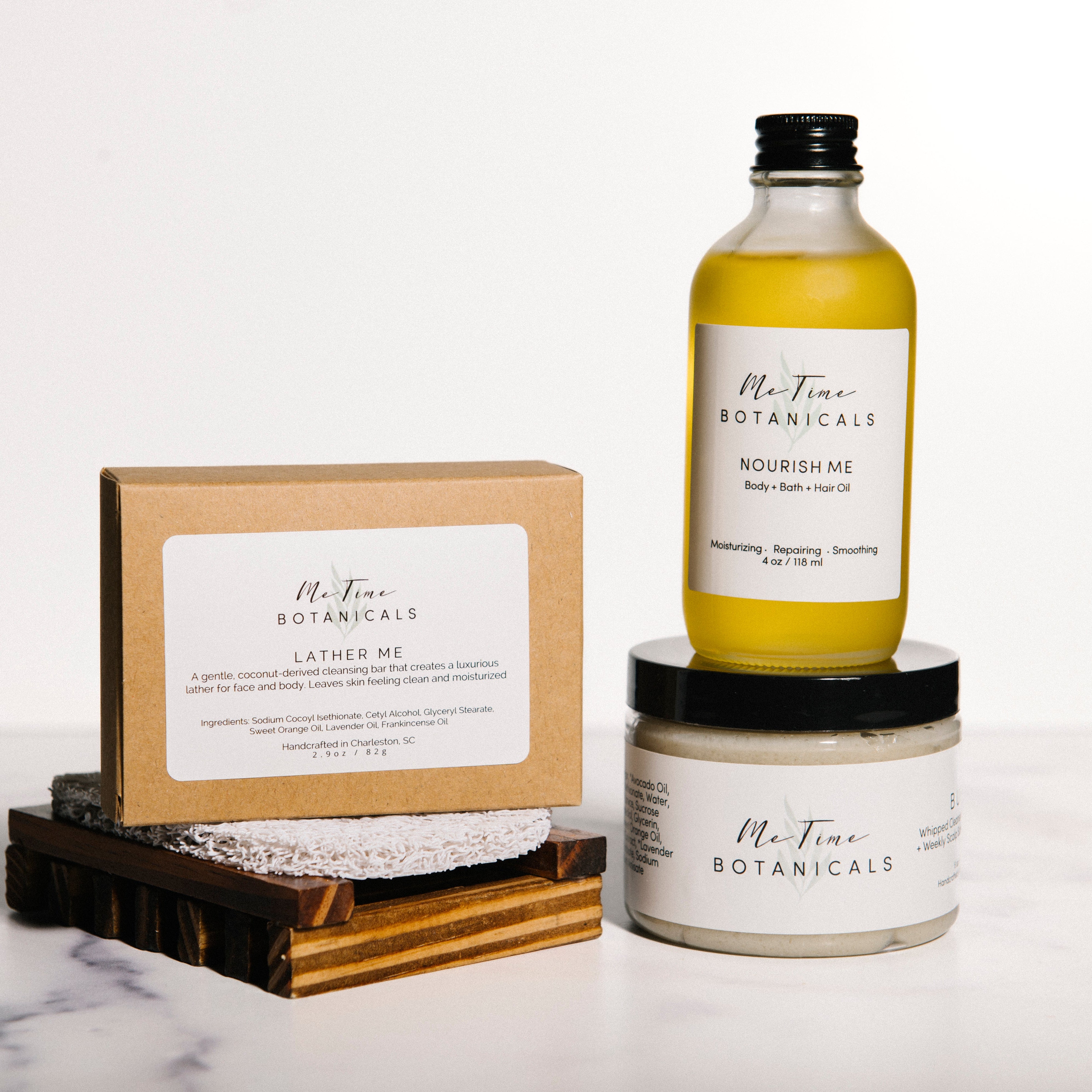 Three Me Time Botanicals bath products arranged on a white marble surface against a white background: a boxed bath salt, a small bottle of oil, and a jar of cream, with the bottle and jar stacked.