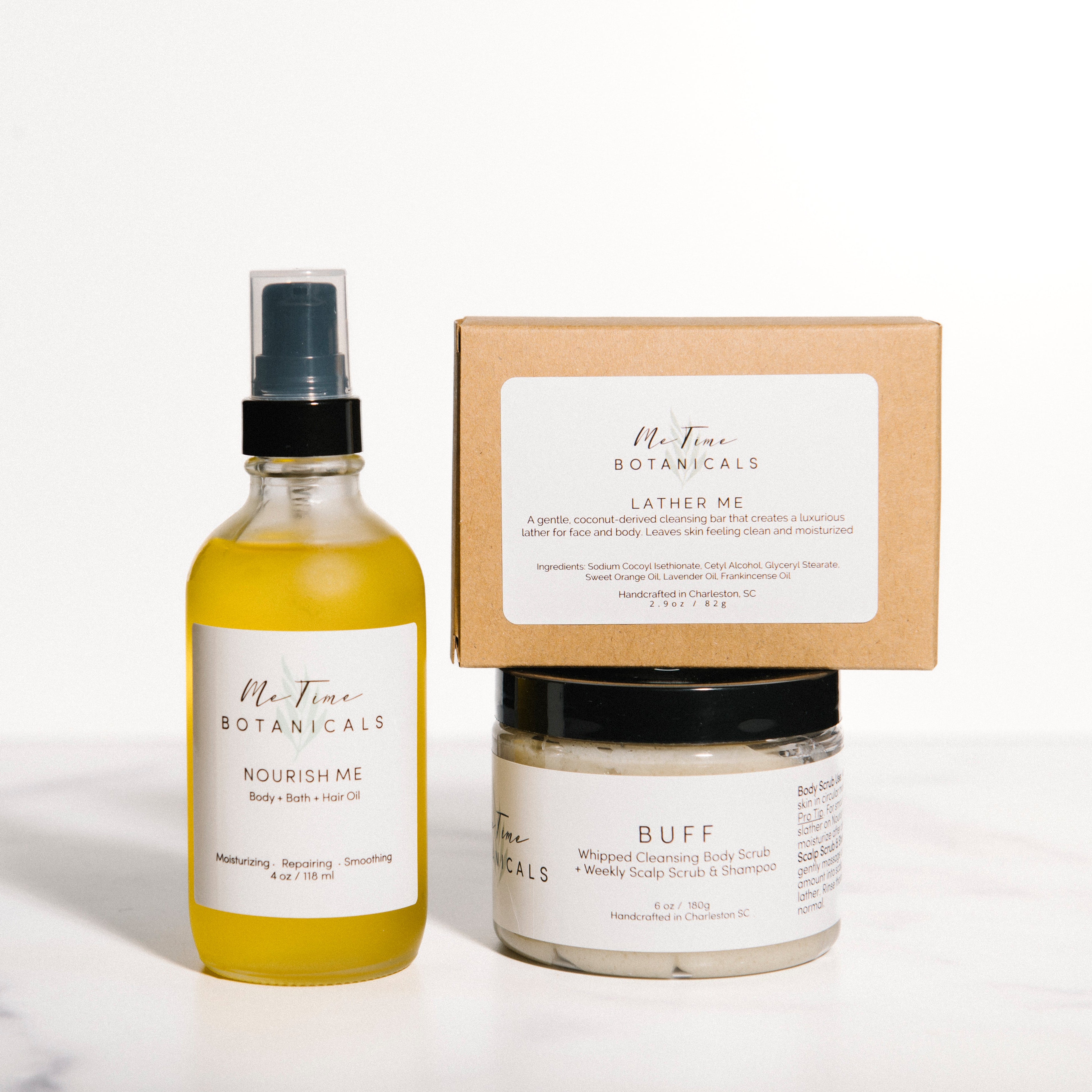 A skincare set from Me Tine Botanicals on a white marble surface against a white background. The set includes a yellow-toned 'Nourish Me' facial toner spray, a white 'Buff' face mask jar, and a kraft paper box labeled 'Lather Me'.