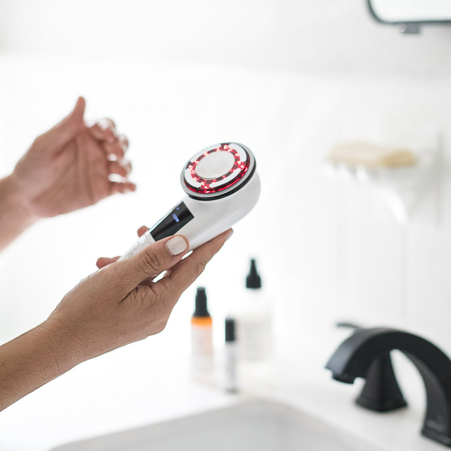 "Hands holding Me Time Botanicals Everything Device with red LED light activated, using microcurrent and  ems showcasing its multifunctional skincare technology, in a modern bathroom setting with skincare products in the background